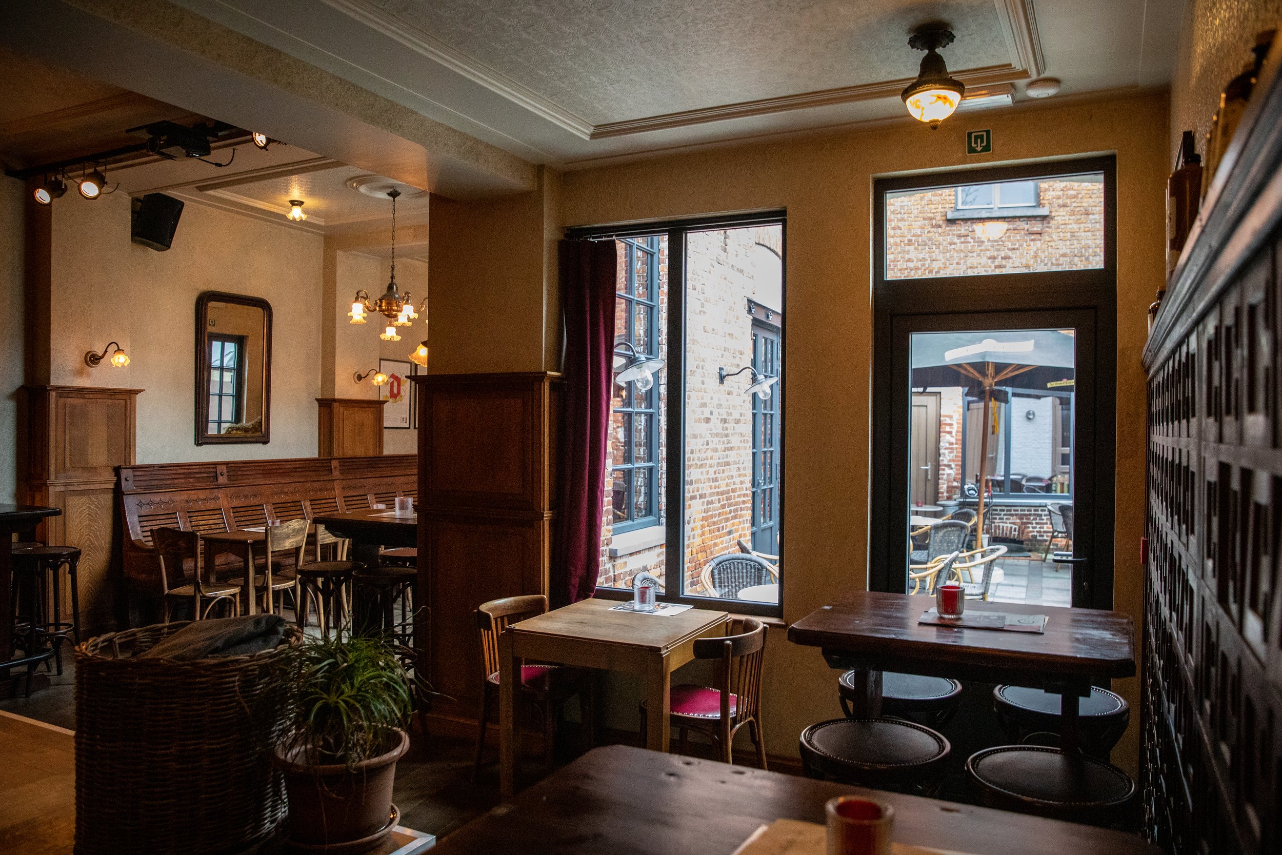 Interieur van een gezellige café met houten meubels en grote ramen die uitzicht bieden op een buitenruimte met terras en parasol.