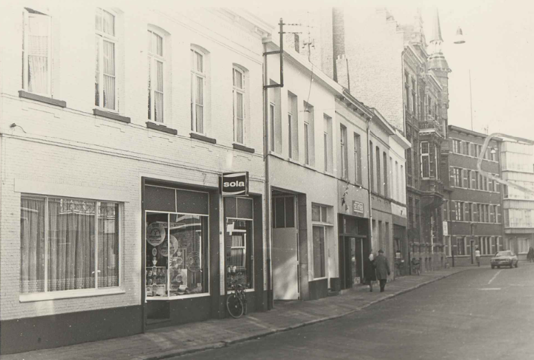 Straatbeeld met winkels, mensen en een auto, zwart-witfoto, vermoedelijke periode mid-20e eeuw.
