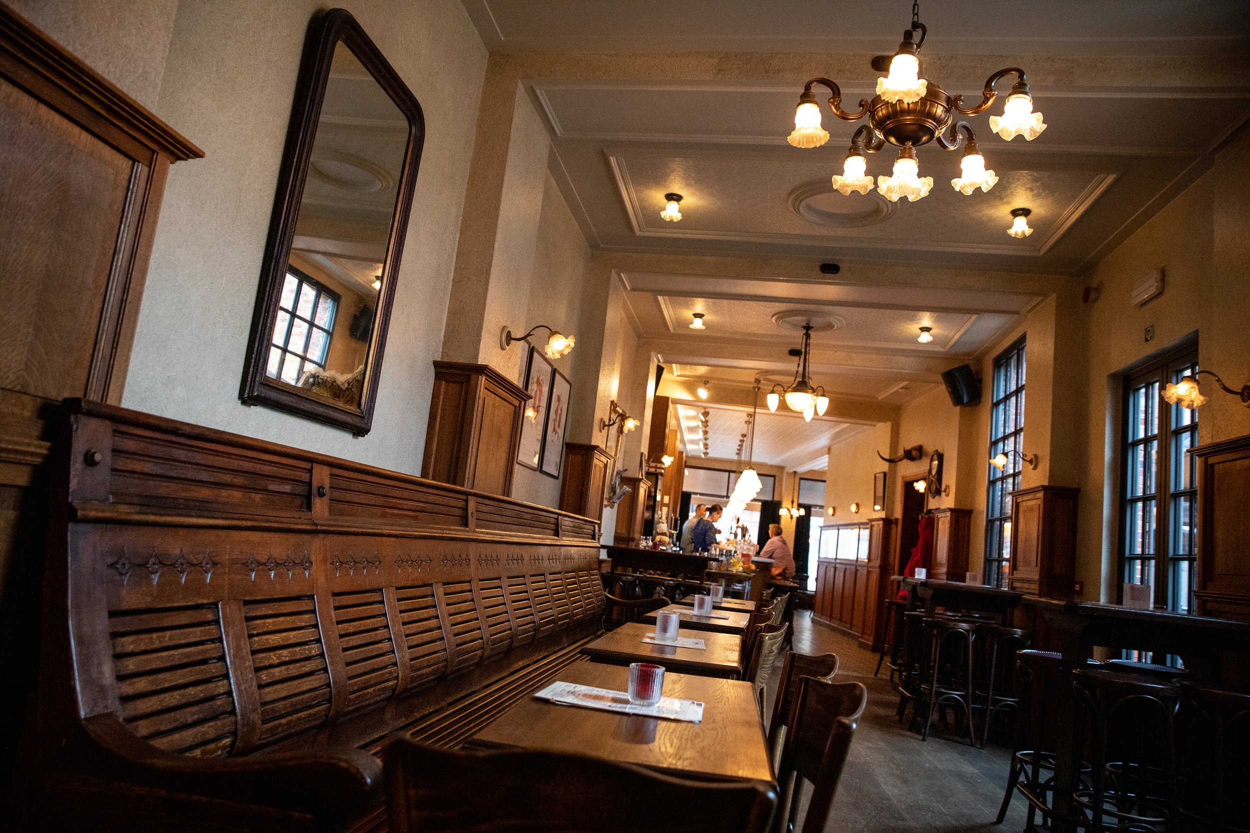 Interieur van een gezellige, traditionele cafe of restaurant met houten meubels, tafeltjes, grote ramen, en vintage plafondlampen.