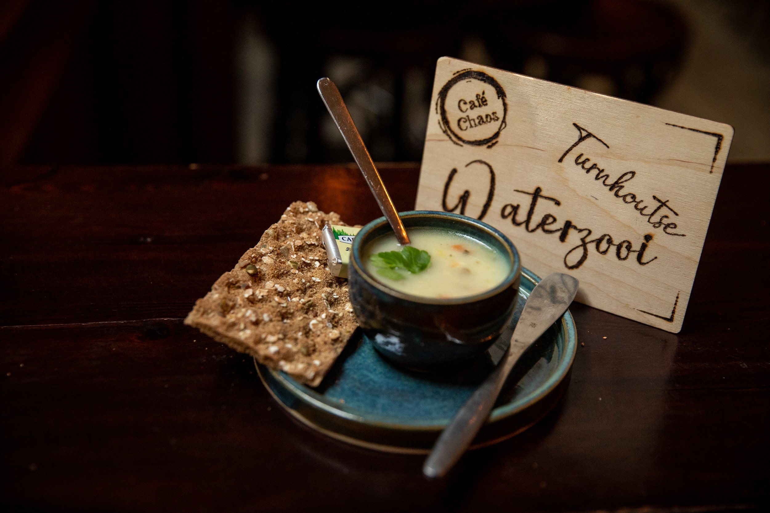 Een kom met soep, een stuk volkorenbrood met zaden, een boterknijper en een houten bord met de woorden 'Trouwhoorsl, Waterzooi' en 'Café Chaos'.