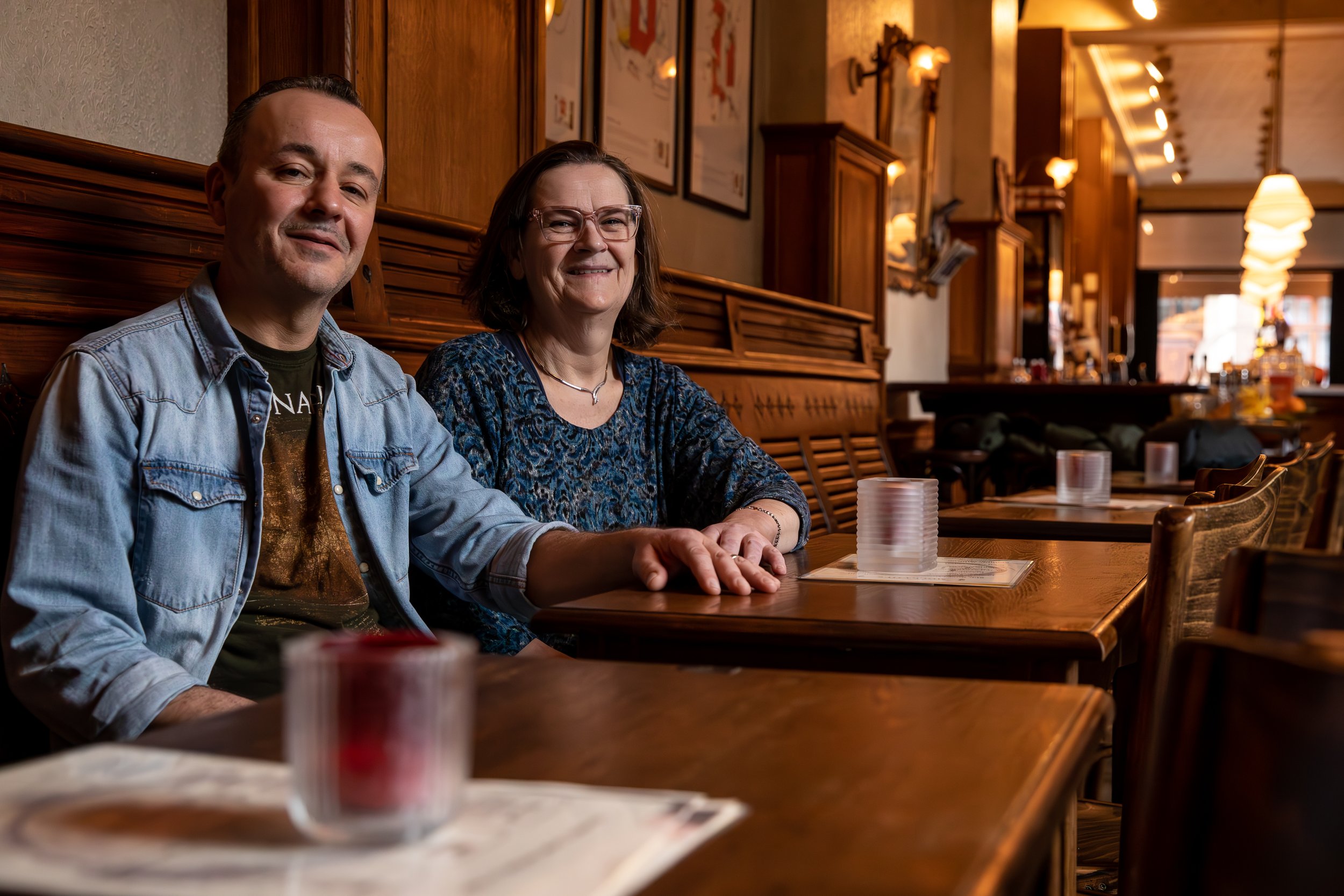 Twee mensen zitten aan een houten tafel in een gezellige restaurant met houten interieur en warme verlichting, lachend en kijkend naar de camera.