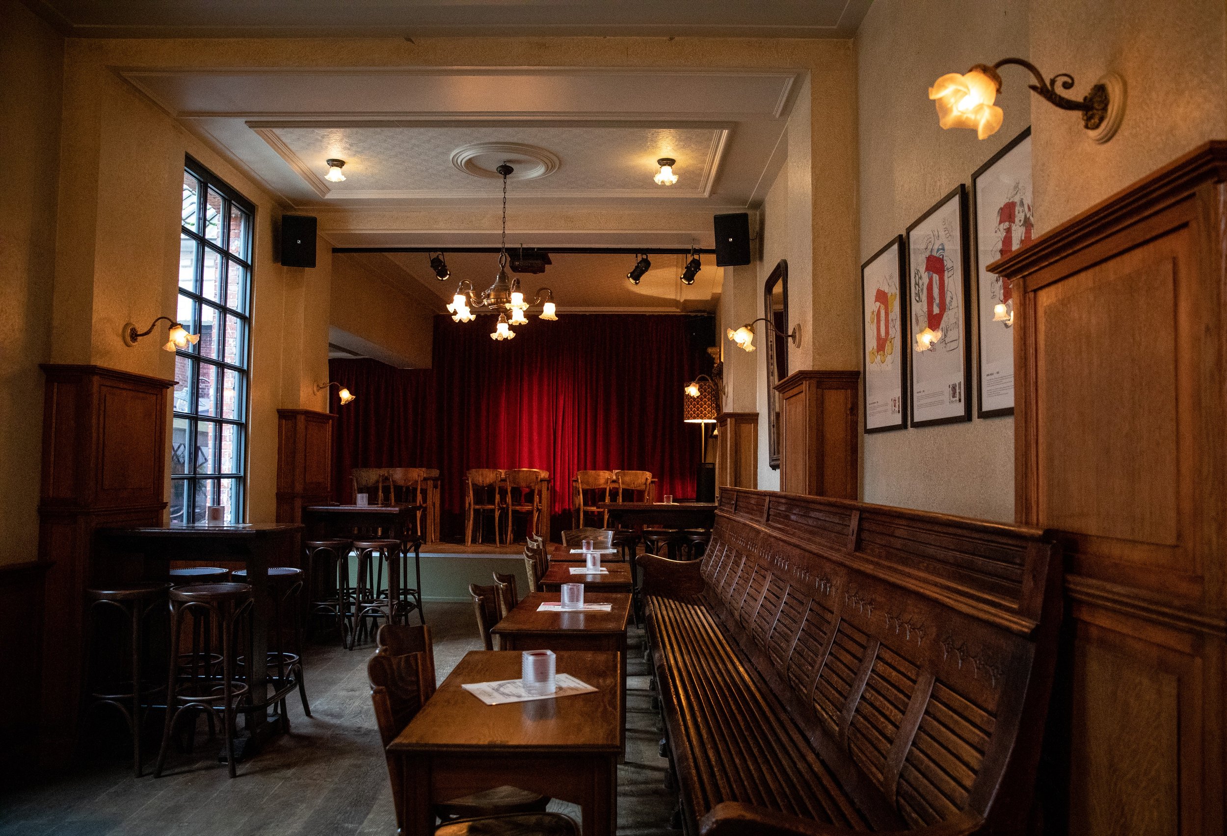 Interieur van een gezellige, donkerverlichte café of kleine concertzaal met houten meubels, een kleine podium met gordijnen, kroonluchters, muurverlichting en betegelde muren.